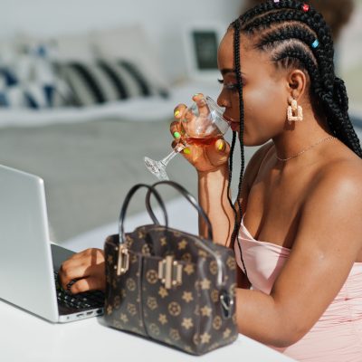 Fashionable photo of young beautiful romantic african american woman with glass of wine, wearing pink dress working with laptop.