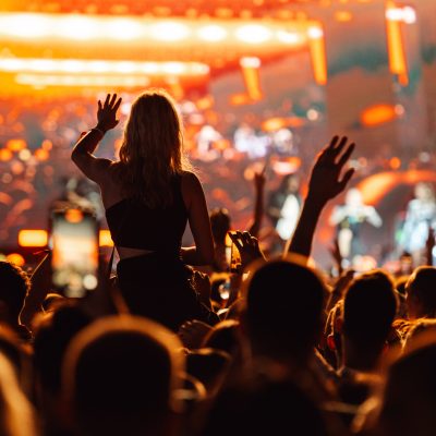 Young woman is sitting on someone's shoulders at a crowded concert, waving one hand in the air while people around her are recording the show on their phones
